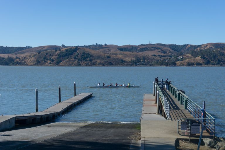 A boat ramp descending to water as rowers in a long sculling shell pass by