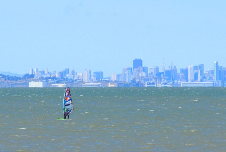 A lone windsurfer on open water with the San Francisco skyline in the background