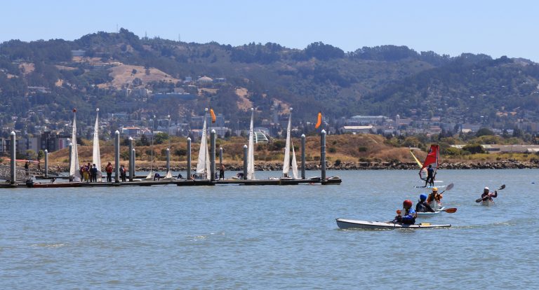 Kayakers paddling on blue water as sailboats and other kayakers float along in the near distance