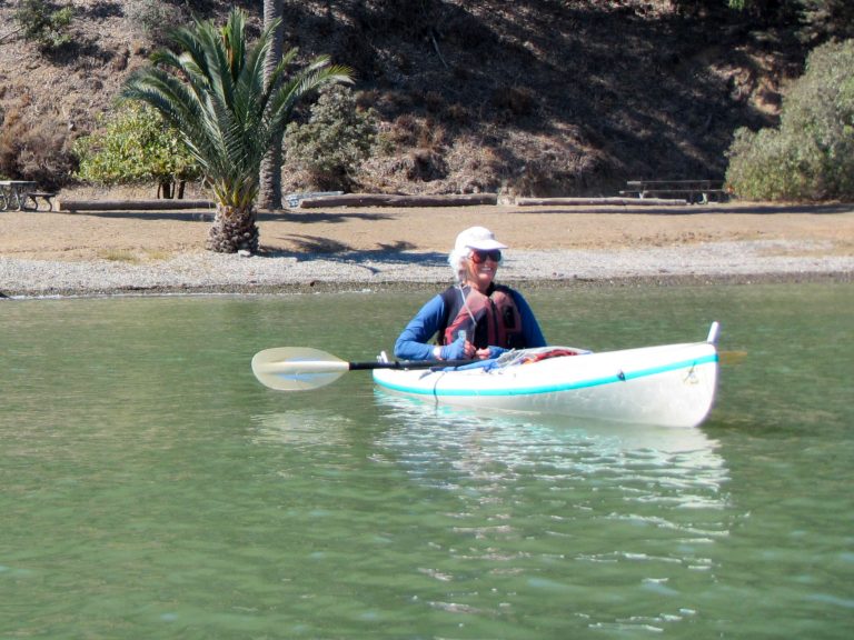 Woman in sunglasses smiling from single kayak, just off the beach