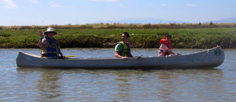 Three people in an aluminum canoe smiling and traveling through a marshway
