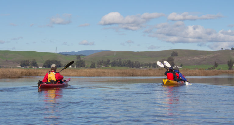 Two kayakers paddling away from the camera, toward a marsh and green hills in distance