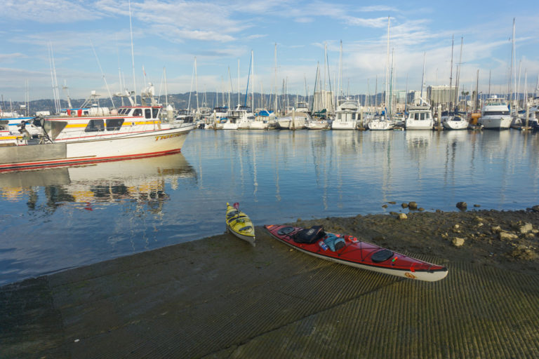 A yellow and red kayak parked on a concrete boat ramp with sailboats moored in the marina