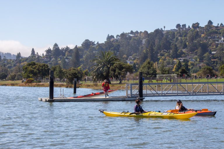 Two single kayakers side-by-side talking while a third person lifts a kayak on the floating dock nearby